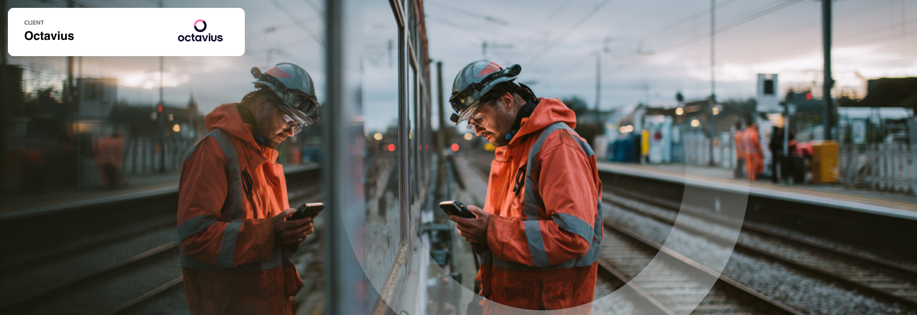Railway worker in high-visibility gear checking a phone, with his reflection visible on a train window at a station.